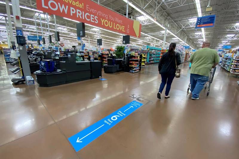 (Top) Red sign with yellow and white text, "Nevada! Everything You Want. LOW PRICES YOU'LL LOVE" Blue sign on floor with white text, "Please keep 6 feet apart" with 2 arrows, one pointing up and one down. Two shoppers, one with black clothes and another with a green shirt and jeans pushing the shopping cart. 