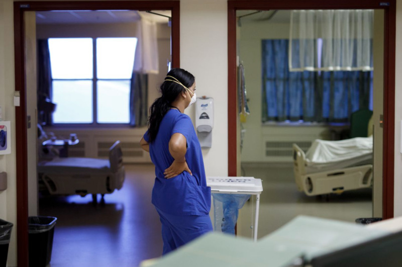 Healthcare worker wears a mask and stands in the hallway of a hospital with her hands on her back.