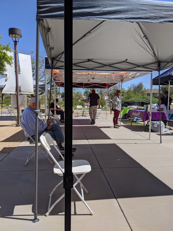 This is a picture of several canopies covering a sidewalk to provide shade for folding chairs. There are several people in the background.  