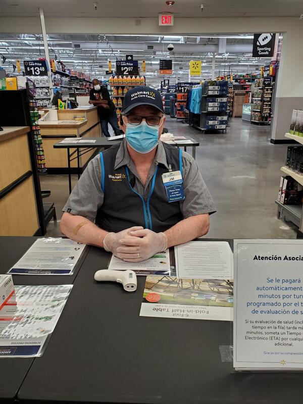 A picture of a Walmart employee wearing a mask and gloves waiting patiently at a table in the store. 