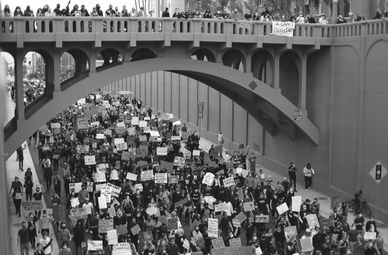 This is a black and white photo of a street that runs underneath a bridge. Both the bridge and the street below are filled with protesters carrying signs related to the Black Lives Matter Movement. 