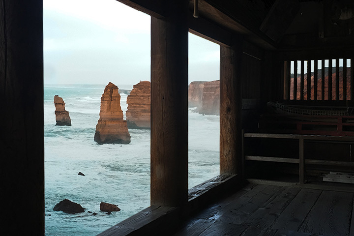 A photo, taken under a wooden covering, of a body of water and breaking waves. Three upright rock columns are scattered in the water as well as cliffs in the background. 