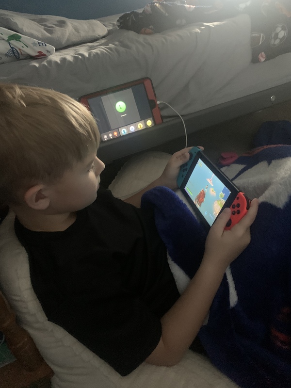 A child sitting next to a bed playing a Nintendo Switch while on a group video call. 
