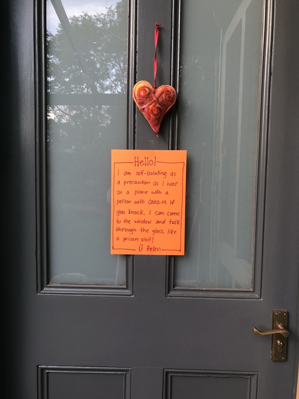 Image of a sign on a door which reads hello, I am self-isolating as a precaution, as I was on a plane with a person with Covid-19. If you knock, I can come to the window and talk through the glass like a prison visit. Helen.