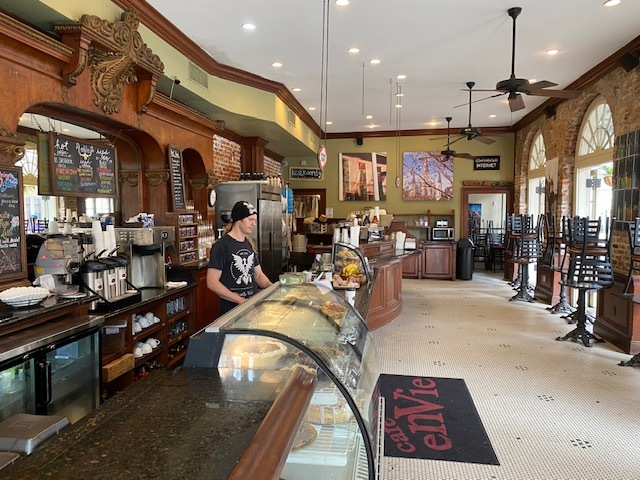 A cafe with no customers. A barista is standing at the register with a black beanie on. All of the black tables have the chairs on top of them showing you are not allowed to sit there. 
