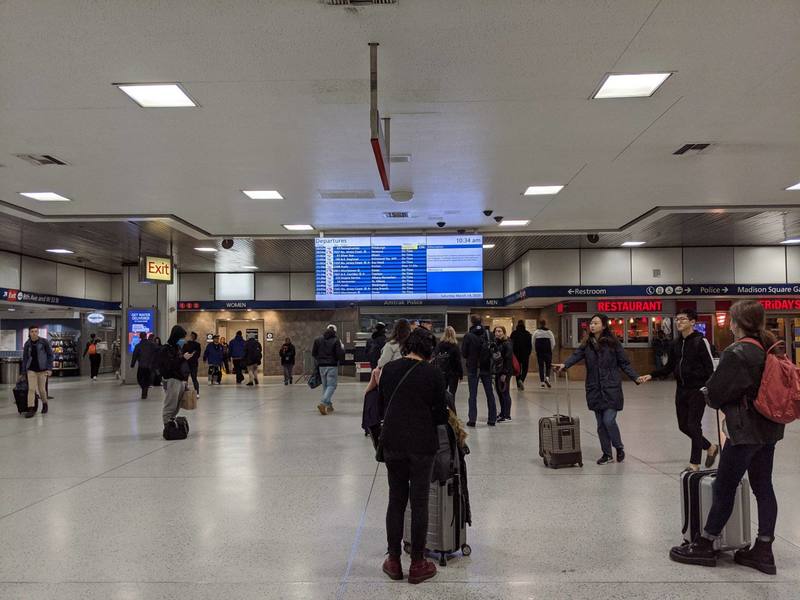 A busy Penn station that has people walking all around with luggage. Near the ceiling is a big screen that has listings of departures.