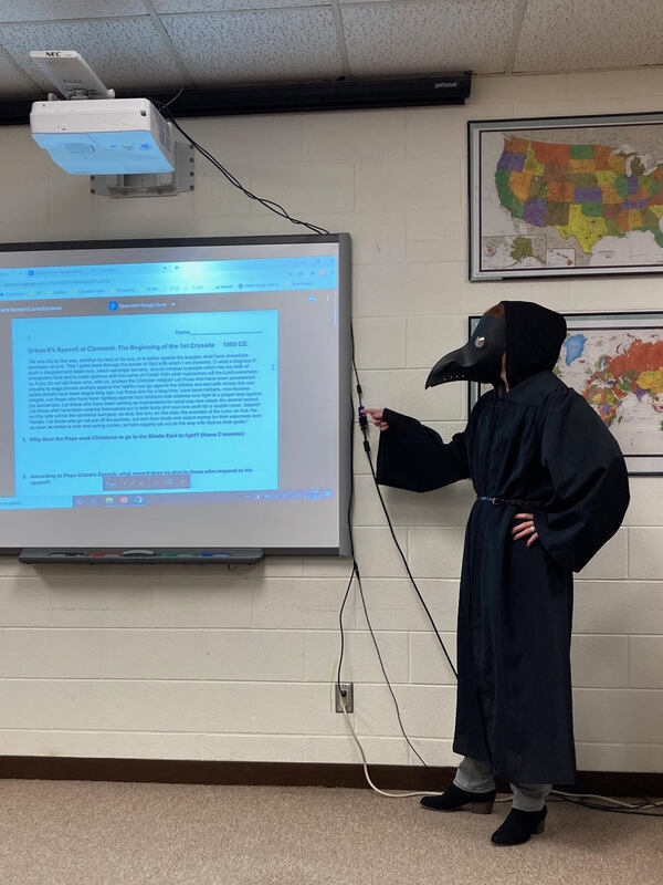 This is a picture of a teacher wearing a plague doctors mask and robe giving a power point lecture in a classroom. World maps can be seen on the wall in the background. 