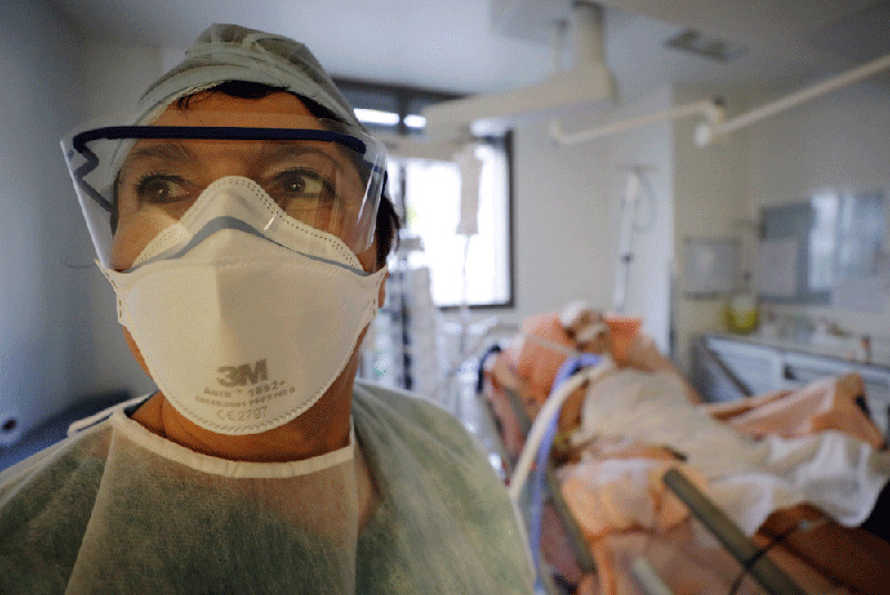 Female medical worker in personal protective equipment in the foreground with patient in hospital bed in the background. 