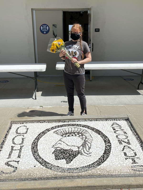 This is a picture of a woman carrying a bouquet of flowers in front of a few collapsible tables and an open door. She is wearing a black face mask, and an APUSH shirt. 