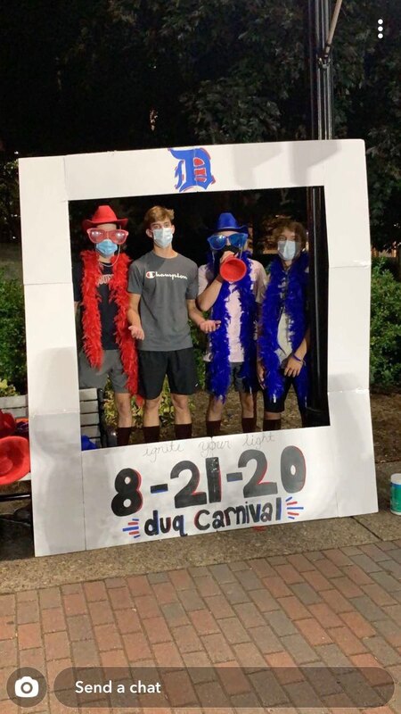 Four college students in masks, hats, and feather boas stand in a photo frame booth at Duq Carnival.