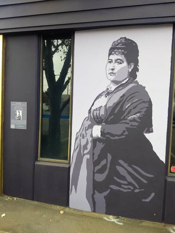 This is a picture taken of a mural that is painted on the side of a building. A woman in a long flowing dress is depicted in black and white. 