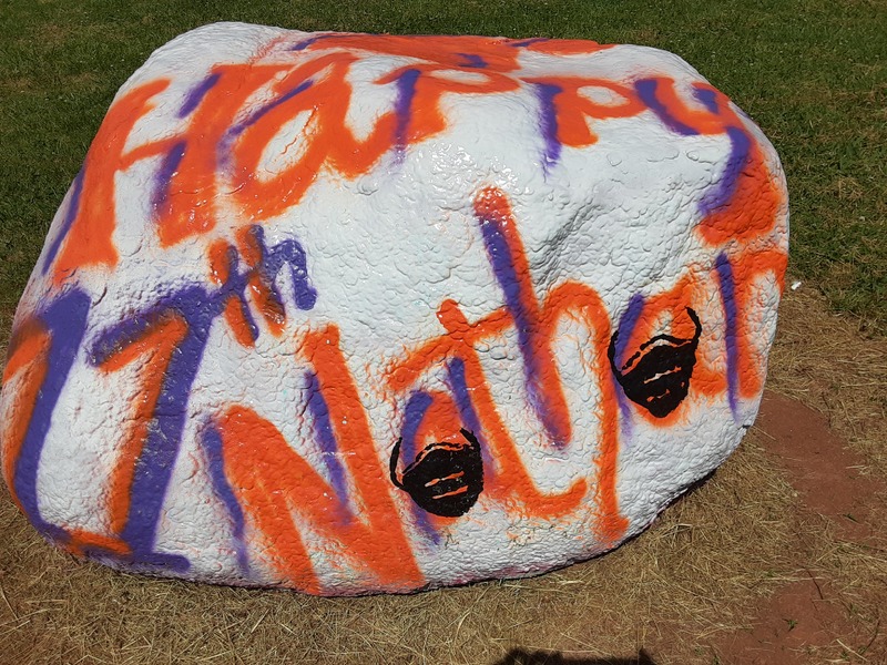 A white rock with the words "Happy 11th Nathan" painted in red and blue on the rock. The A's in "Nathan" have black face masks painted on top of them.