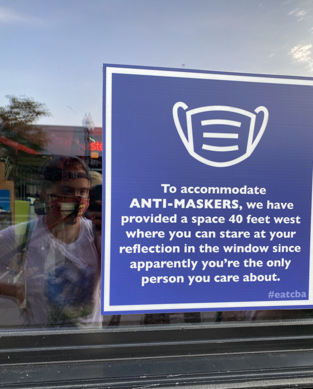 This is a picture taken of a sign placed on the window of a business. The sign has an image of a face mask on it and reads: "To accommodate ANTI-MASKERS, we have provided a space 40 feet west where you can stare at your reflection in the window since apparently you're the only person you care about."