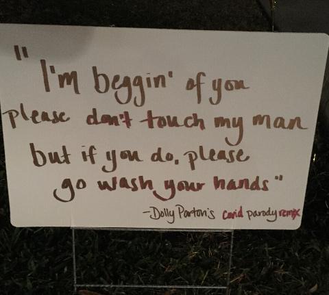 This is a picture of a white sign placed in a lawn which reads: "I'm beggin' of you please don't touch my man, but if you do, please go wash your hands. -Dolly Parton's COVID parody remix."