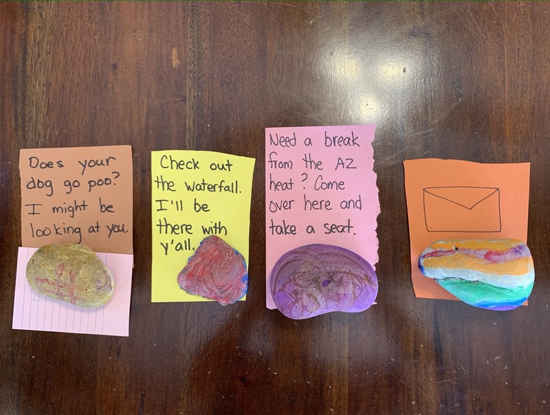 Four colorful rocks are sitting on pieces of paper that have clues written on them and are all placed on a wooden table. 