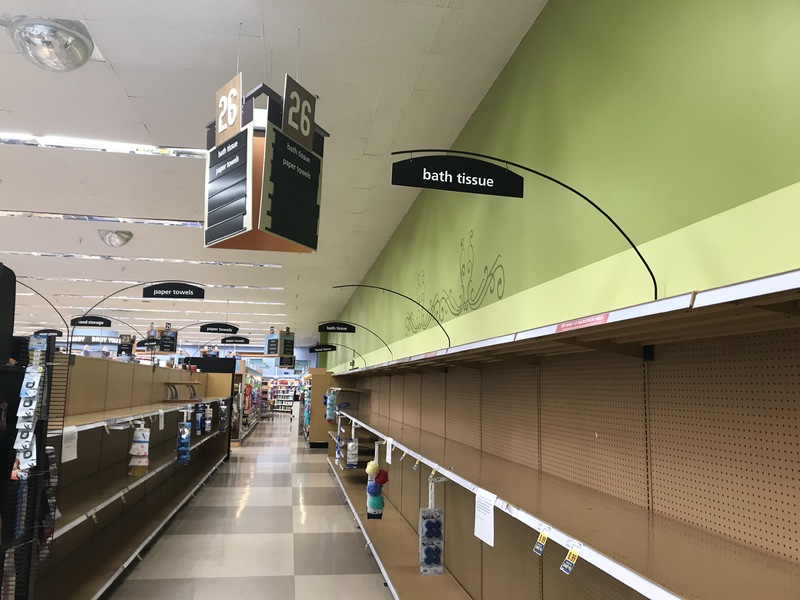 A supermarket that has green walls with checkered beige flooring. There are rows of empty shelves. Above the shelves is a black sign with white text that says "bath tissue". Hanging from the ceiling above the isle has a triangle shaped brown and beige sign that signs 26 with beige lettering. 