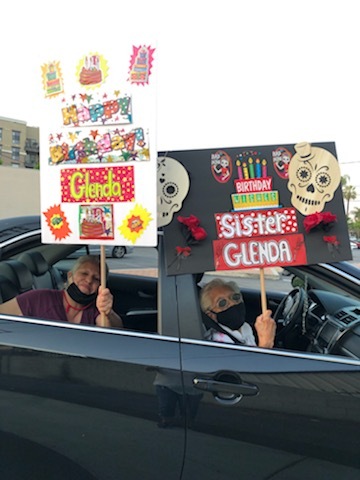 This is a picture taken of two elderly women wearing face masks and carrying signs while riding in a car with the windows down. They appear to be celebrating another persons birthday while adhering to social distancing standards. The colorful signs they are holding read "Happy Birthday Glenda."