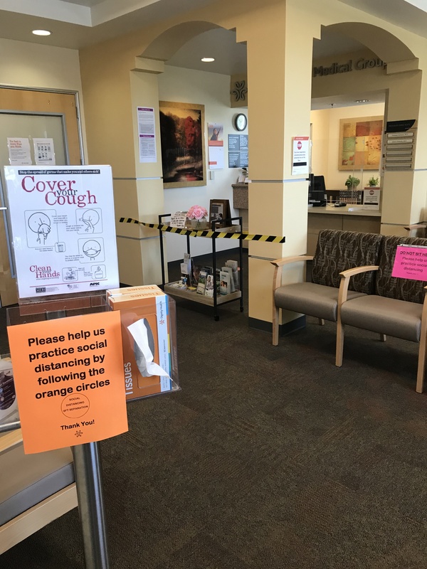 Signs in the waiting room at a medical center instructing visitors to cover their cough and to please help us practice social distancing by following the orange circles on the floor indicating six feet apart. 