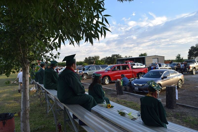 High school graduates sit socially distanced on bleachers outdoors while all attendees watch from their vehicles.
