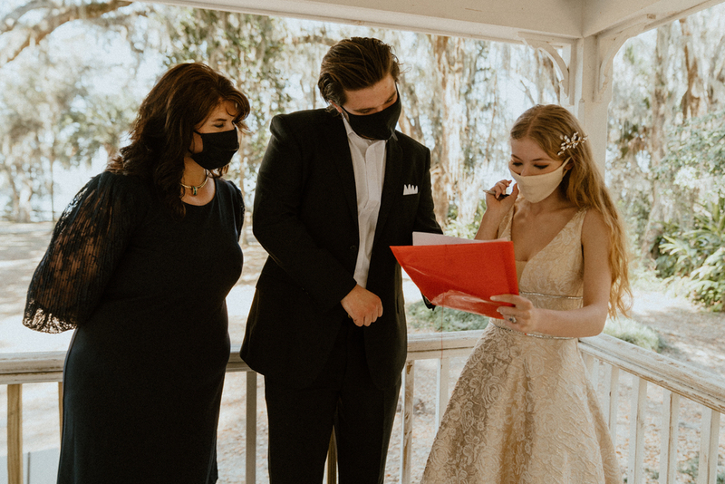 This is a picture of three people dressed in formal attire standing outside on a porch. All are wearing face masks. The woman in the photo seems to be wearing a wedding dress, while the man seems to be the groom. 