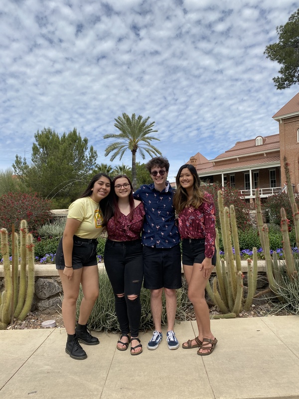 Three girls and a boy standing outside in front of cacti and palm trees, posing together for the photo.