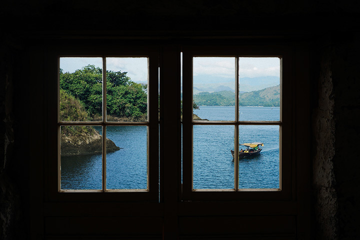 A photo taken through a window overlooking a body of water. A boat is in the water surrounded by steep hills.
