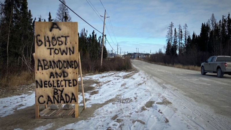 This is a picture taken of a road, flanked by trees. A wooden sign on the side of the road written on with spray paint reads "A ghost town abandoned and neglected by Canada."