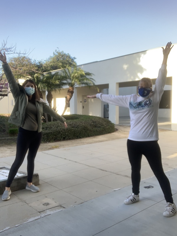 This is a picture of two women wearing face masks, raising their arms and posing for the picture. They are both wearing black pants, and the woman on the left is wearing a green coat and shirt. The woman on the right wears a white long sleeved shirt and glasses. 