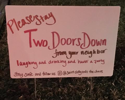 This is a picture of a white sign stuck in someone's lawn which reads: "Please stay two doors down from your neighbor, laughing and drinking and havin' a party. Stay safe and follow us @Saintdollyendsthechaos."