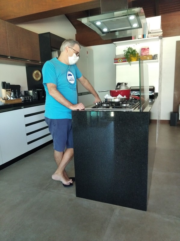A man is standing at an island in front of a stove in the kitchen. The man is wearing a light blue shirt with blue shorts and he is wearing a white face mask. 