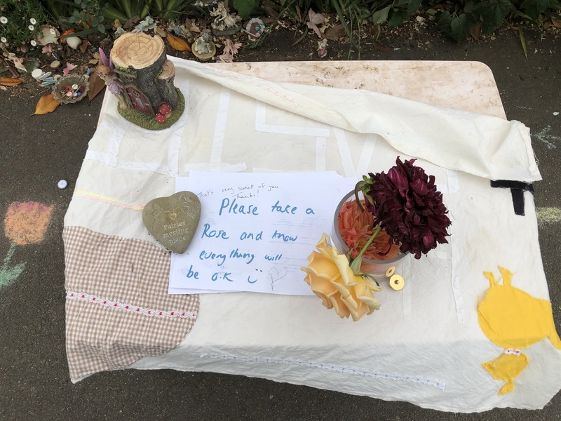 A table with a cloth over it. There's a vase with various flowers in it. There's a paper sign on top of the table that says: Please take a Rose and know everything will be ok (smiley face).