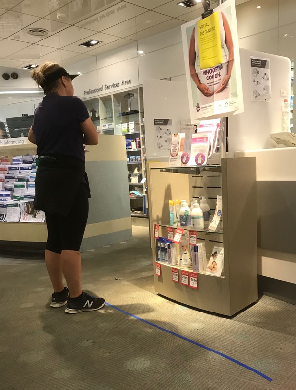 A woman in a pharmacy dressed in all black workout gear wearing a visor and is waiting in line. On the floor is a 6 feet piece of blue tape, measuring social distancing. 