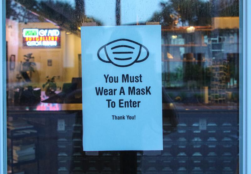 This is a picture of a blue sign that is taped to a window, which reads: "You Must Wear A Mask to Enter. Thank you!" The image of a face mask is also present on the sign. 