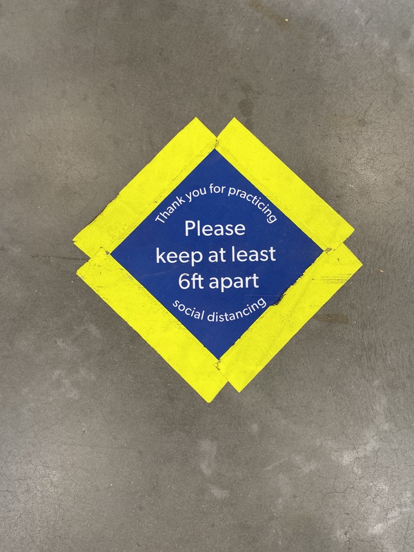 "Blue diamond shape outlined with bright yellow tape on the floor of Sam's Club. Text inside the diamond reads, "Thank you for practicing social distancing. Please keep at least 6ft apart."