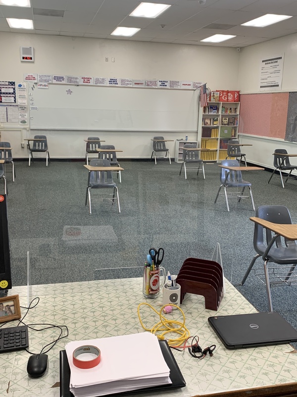 This is a picture of a classroom taken from the perspective of the teachers desk. There are several desks in the room that are spaced far apart, as well as a bookshelf and whiteboard in the back of the room. 