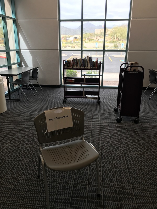 The corner of a library with windows. There are two book carts with books. There is a chair with a paper sign on it that says: Day 1 Quarantine. 