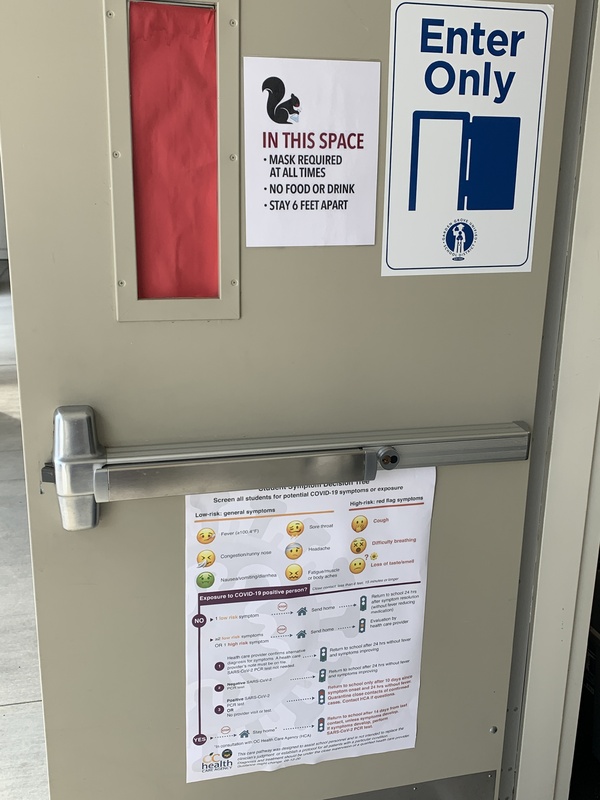 This is a picture of a classroom door that has several signs attached to it. The first one reads "In this space: Mask required at all times, no food or drink, stay 6 feet apart." The second message reads "Enter only" followed by a diagram of a door swinging open. A third message on the bottom of the door appears to depict a chart which showcases various information about COVID-19 symptoms. 