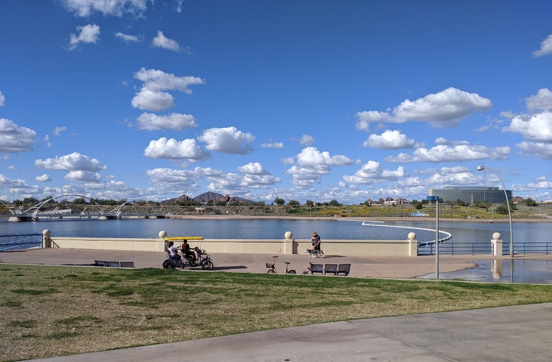 A park that overlooks a body of water, the skies are blue with fluffy clouds. On the right on the sidewalk is an old couple that is walking their dog that is a husky. On the left of the sidewalk is three people sitting in a black pedal bike that has a yellow covering on top. 