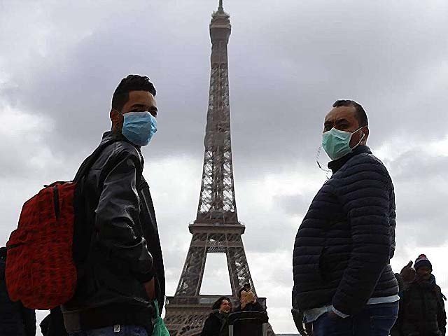 Two people are standing near each other with face masks on. Behind the the two men in the distance is the Eiffel Tower in Paris, France. 