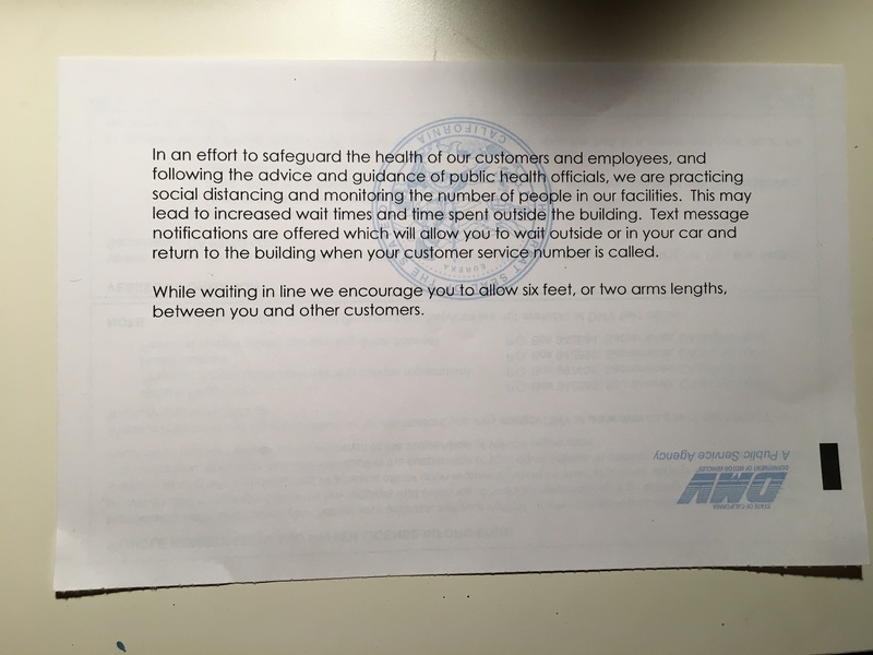 A paper information flyer from the Department of Motor Vehicles taped to a white wall saying they are practicing social distancing and limited number of people inside of their building.  