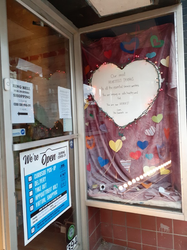 This is a picture taken of a storefront window, where several signs are present. One sign lists curbside pickup, appointment only, and virtual shopping as the only options to purchase items from the store. A paper heart in the window reads: "Our most heartfelt thanks to all the essential service workers. You are keeping us safe, healthy, and fed. You are our heroes! Love the Daniels xoxo."