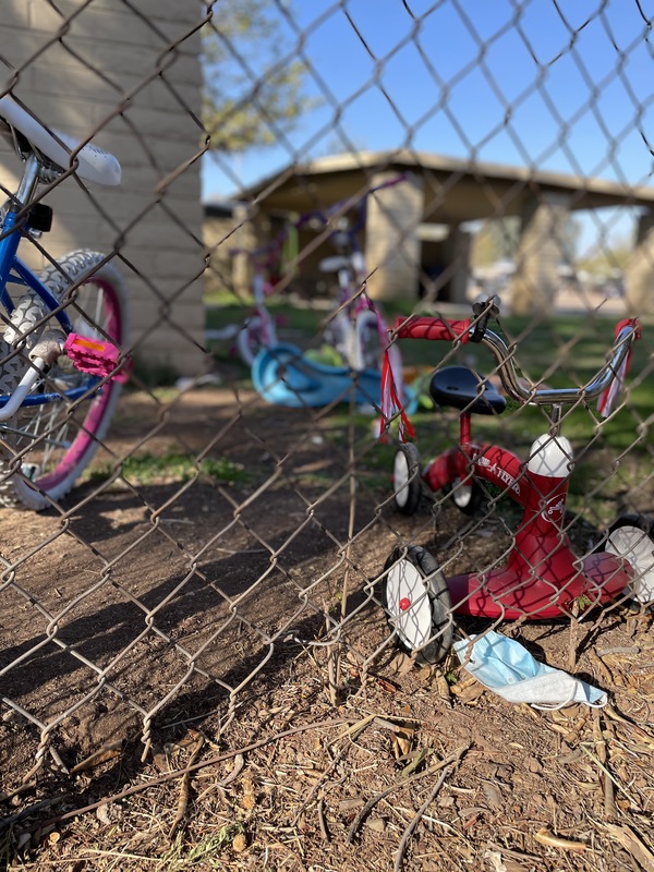 This is a picture of a discarded face mask resting next to a fence. Children's bicycles and trikes are leaned against the other side of the fence.  