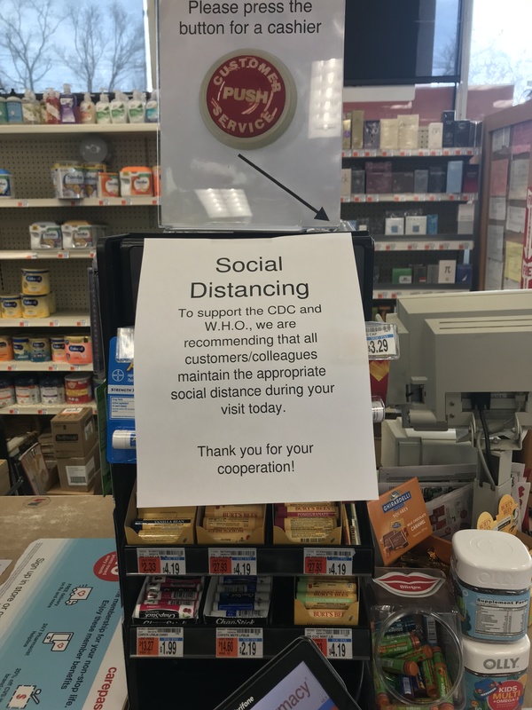 A register with a sign on it that says: Please press the button for a cashier and has an arrow pointing to the button. Below the sign is a paper sign that says: Social Distancing to support the CDC and W.H.O., we are recommending that all customers/colleagues maintain the appropriate social distance during your visit today. Thank you for your cooperation! 