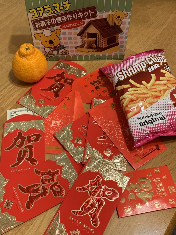 Chinese New Year meal and celebration.  Red envelopes on a wooden table with a mandarin orange, shrimp chips, and a cookie house kit. 