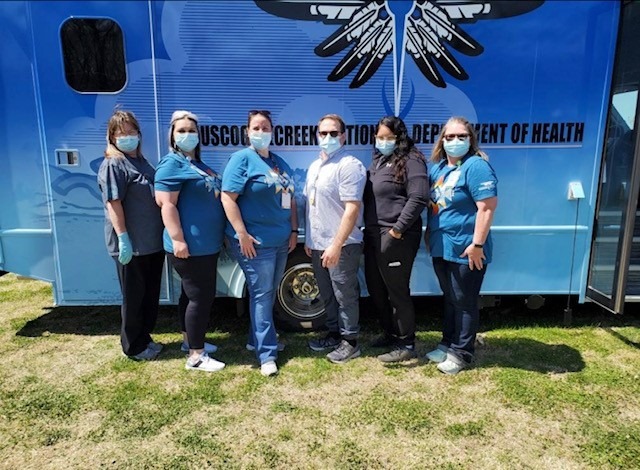 This is a picture taken of six healthcare workers wearing masks posing in front of a bus parked in a grassy area. The bus is painted blue, with the words Muscogee (Creek) Nation Department of Health on the side. 