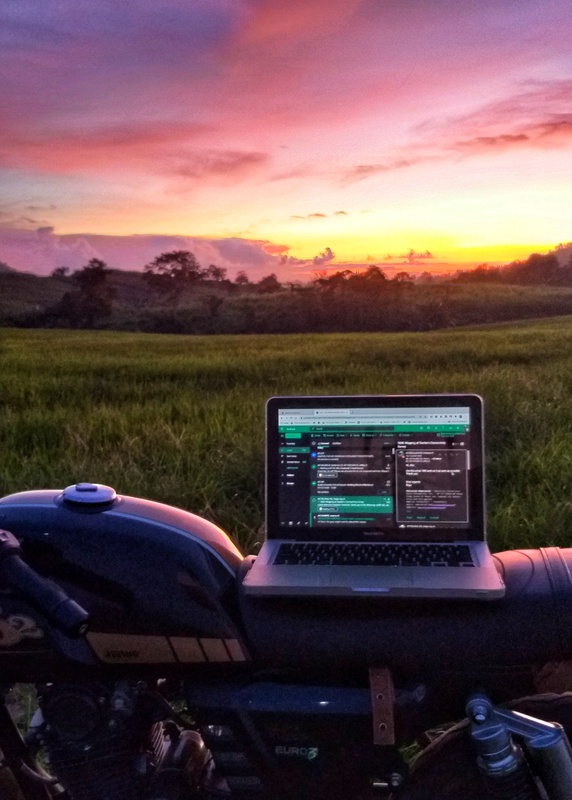 A photograph of a laptop on top of a motorcycle in front of sunset behind rolling hills. 