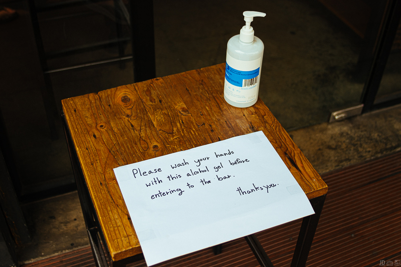 A wooden stool has a paper sign with written sharpie on it that says: Please wash your hands with this alcohol gel before entering the bar. Thanks you. There is a bottle of hand sanitizer on the right upper corner of the wooden stool. 