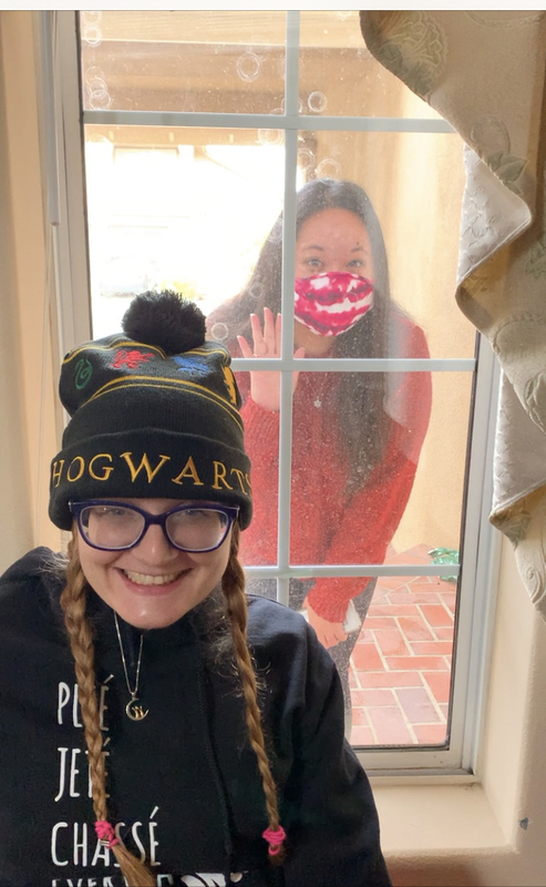 Girl with glasses and hat stands in front of a French door, behind which is a masked woman waving through the glass.