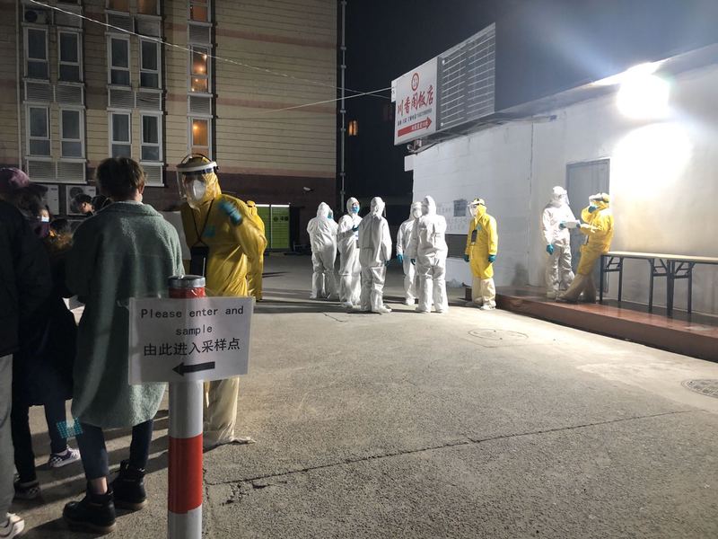 People in yellow or white hazmat suits with people waiting to be Covid tested. 