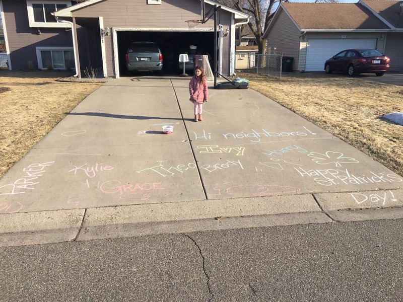 Picture of a young girl in her drive way in front of messages in chalk reading "Hi Neighbors" and "Happy Saint Patrick's Day". 